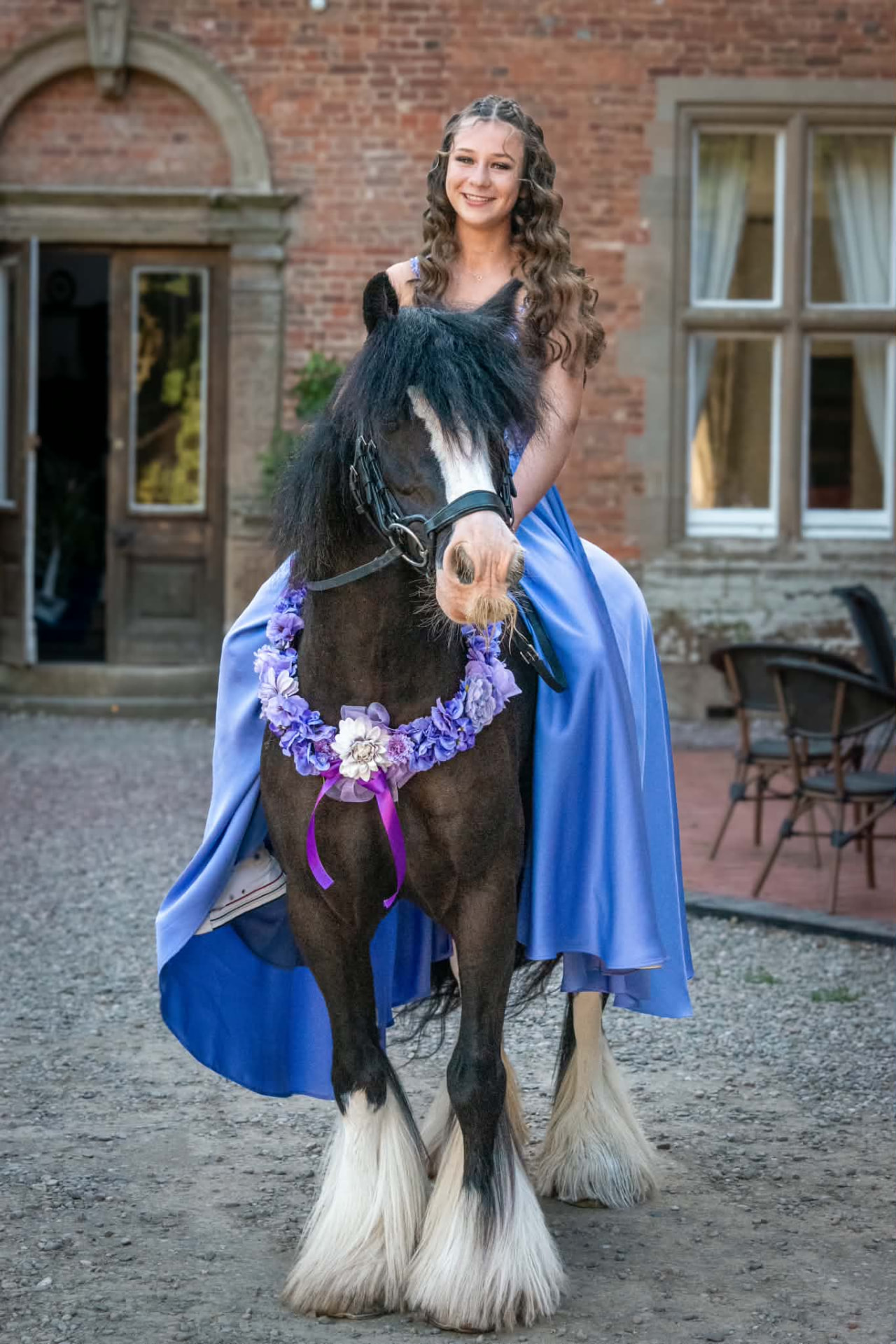 Girl in a blue gown seated on a black and white cob outside a brick house