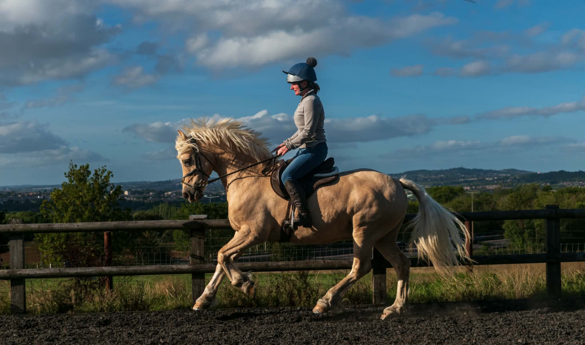Rider on a palomino pony trotting along an outdoor arena against a blue sky